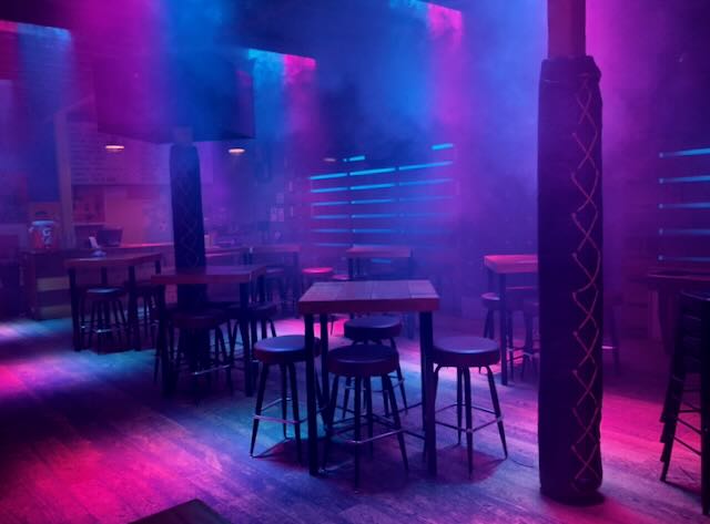 Bar interior bathed in magenta and blue haze, rope-wrapped column anchoring rows of high-tops and stools.