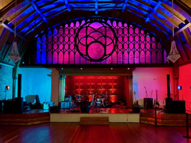 Concert hall stage with a stained-glass rose window, drum kit and amps lit in saturated red beneath cool blue uplight.