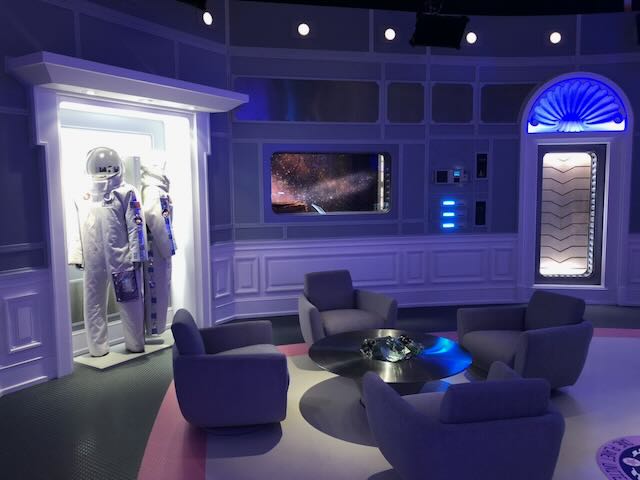 Sci-fi lounge set — astronaut suit, deep-space porthole, and curved seating washed in cool blue and violet.