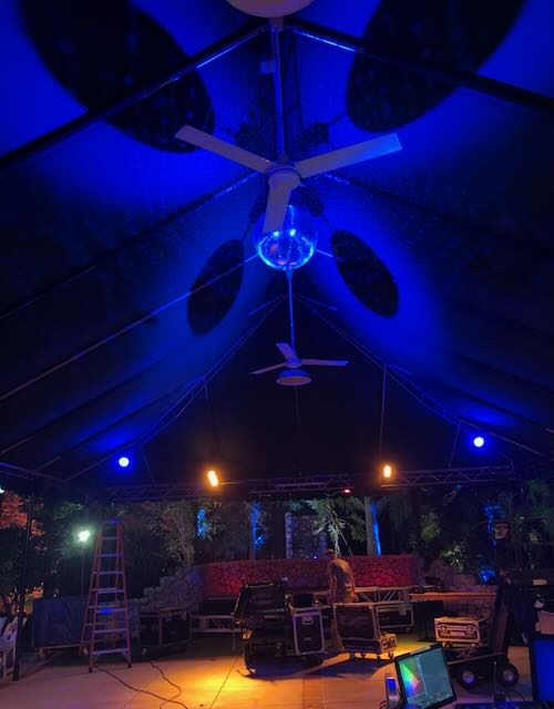 Tented outdoor venue under deep blue uplight, with practical ceiling fans and warm pool lighting on the stage.