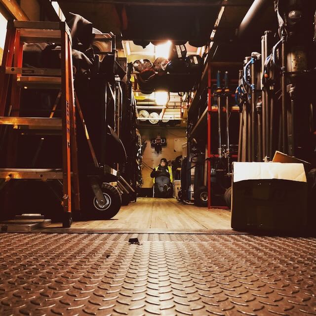 Backstage corridor stacked with ladders, stands, and road cases — a crew member silhouetted at the far end.