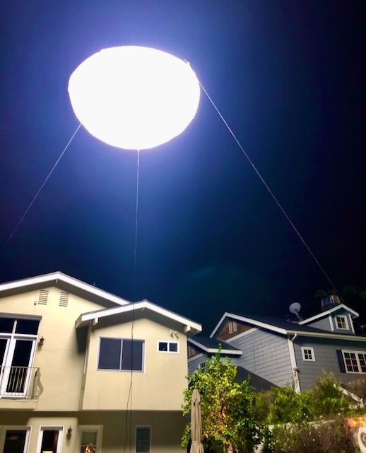 Helium balloon light glowing above a residential rooftop at night, used as a soft moonlight source.